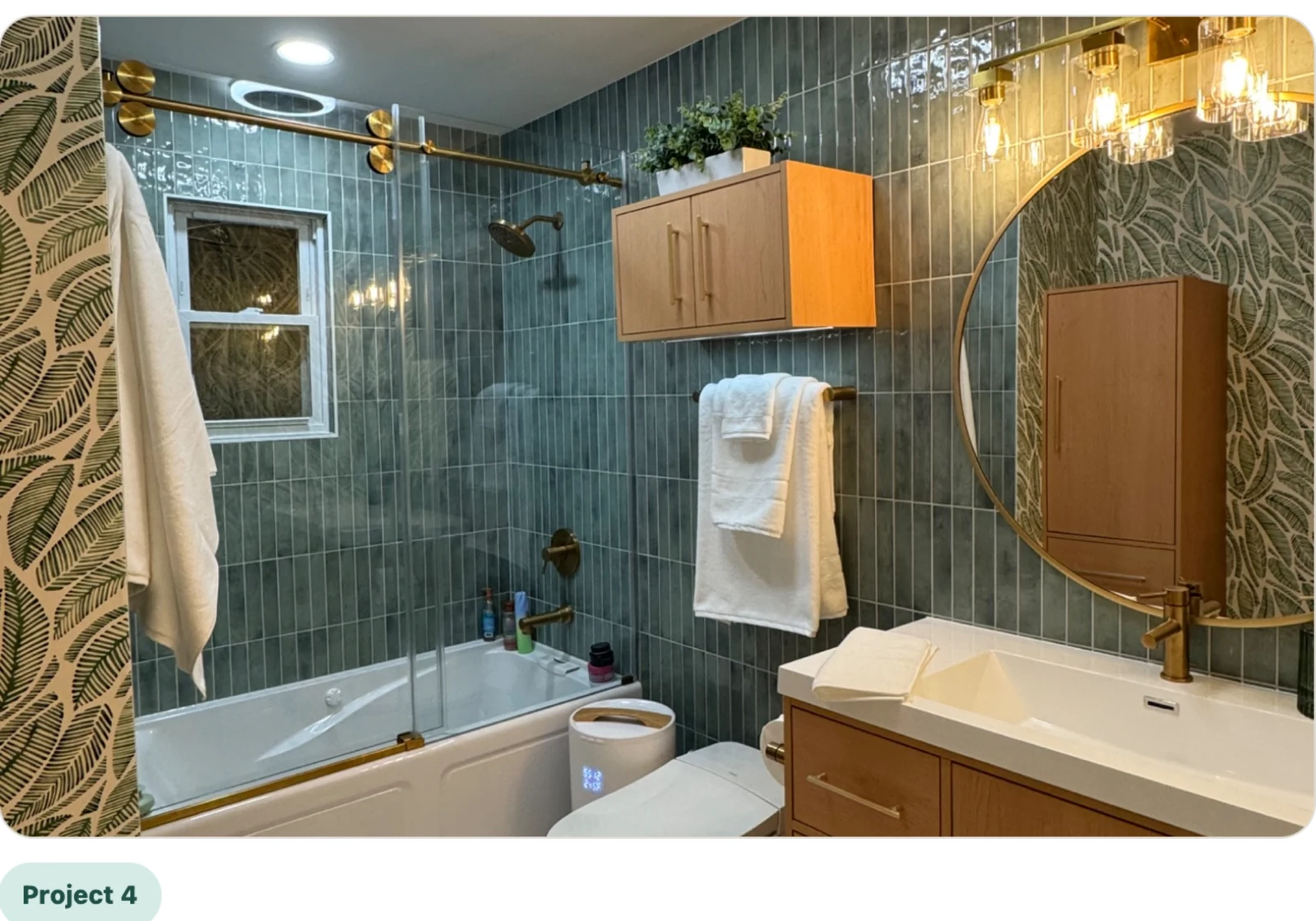 Finished bathroom update with green tile, glass tub door, warm wood vanity, and brass fixtures