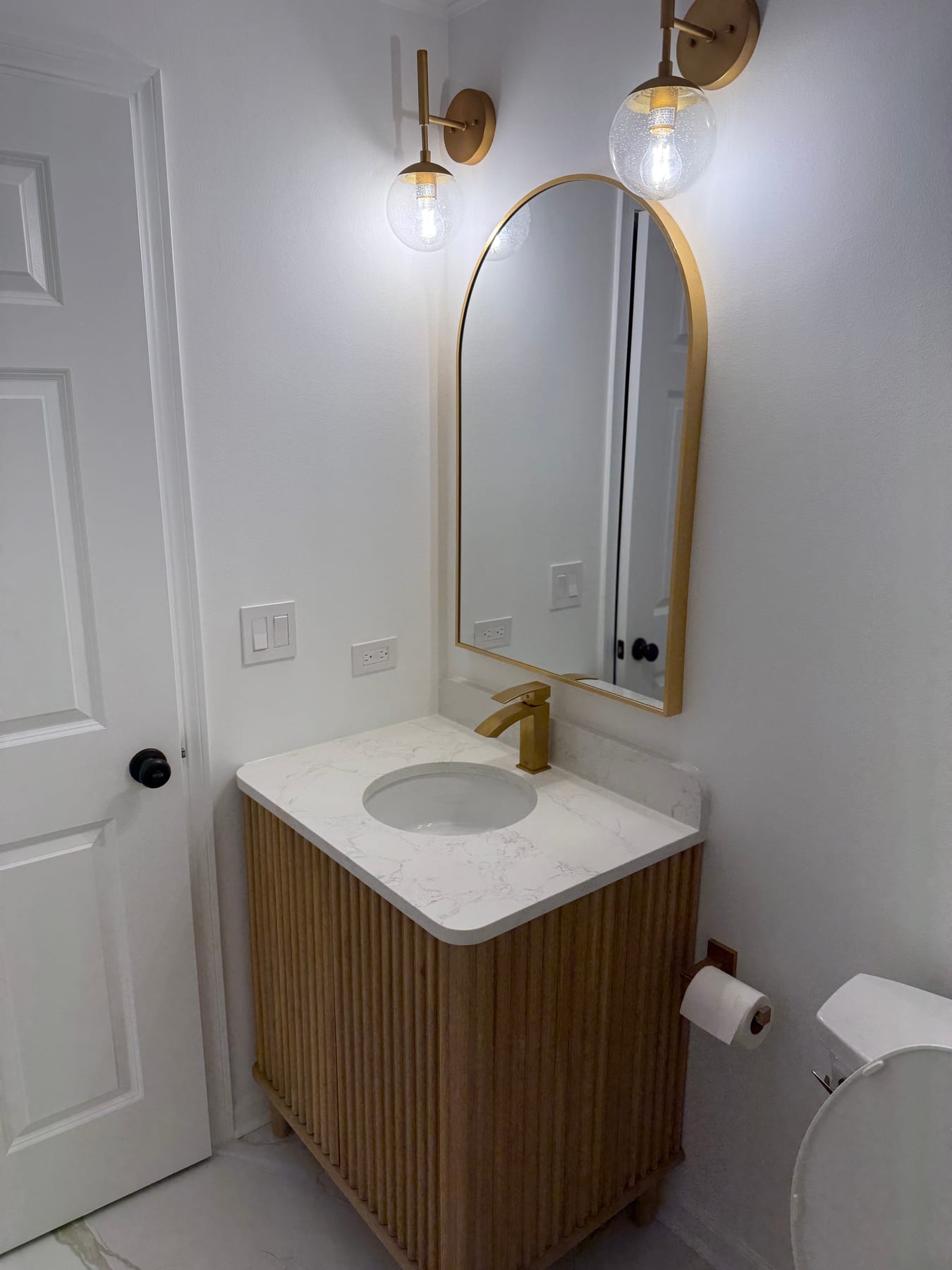 Wood bathroom vanity with gold faucet, arched mirror, and matching wall sconces